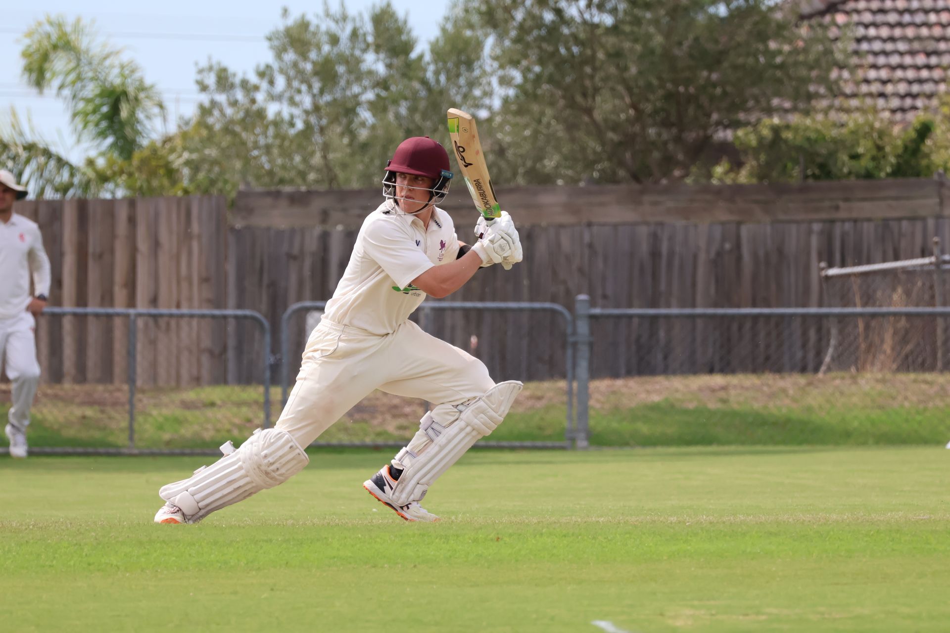 Cricket batter in white runs across a grassy field with bat raised, wooden fence and trees in background