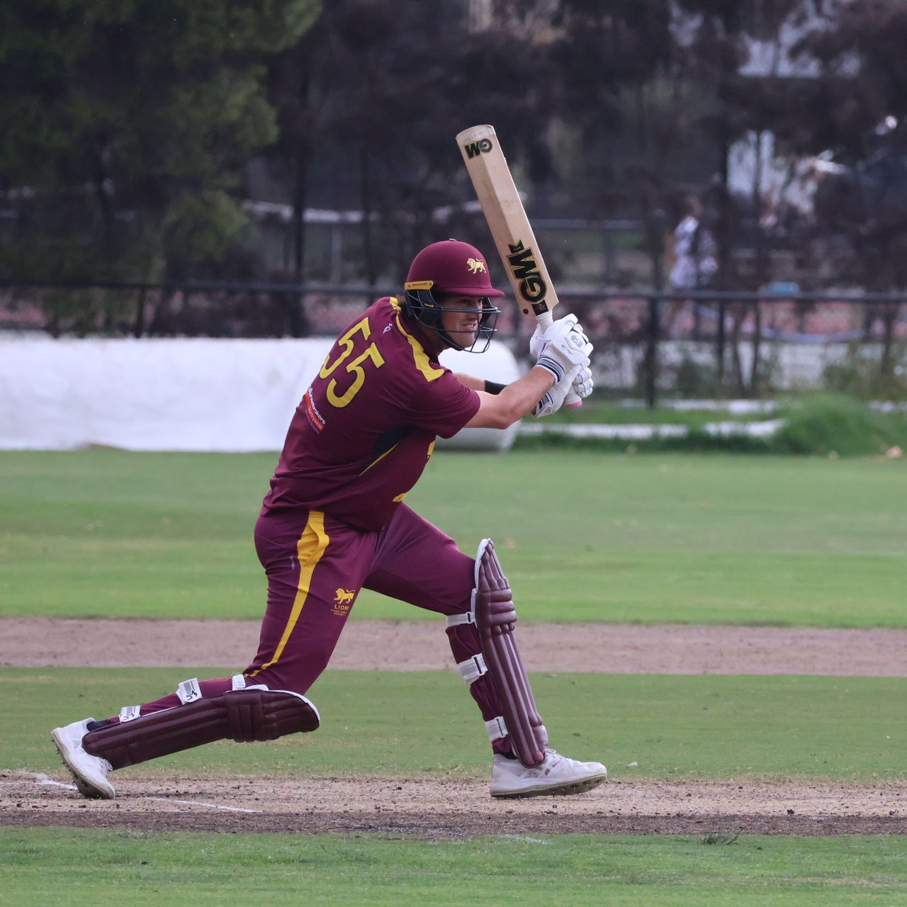 Cricket batter in maroon uniform swings a bat on a grassy field.