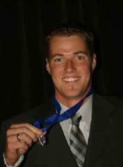 Smiling person in a suit holding a medal ribbon against a dark background