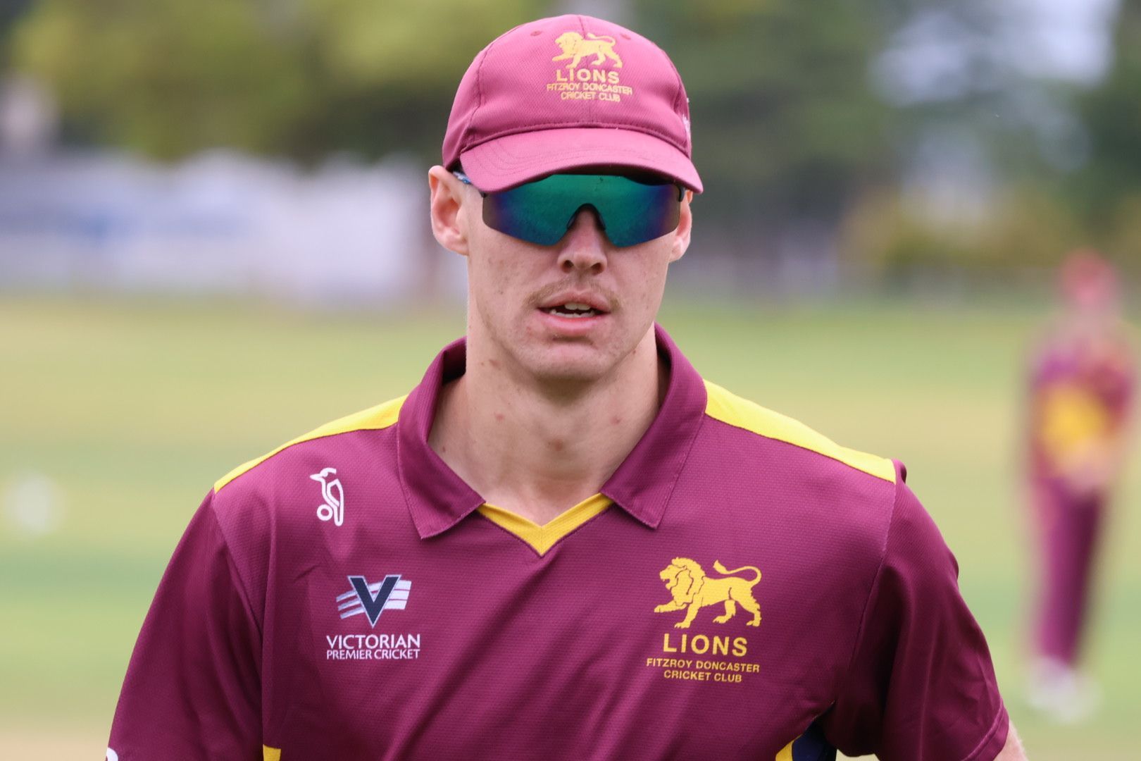 Cricket player in maroon cap and jersey with reflective sunglasses on a field
