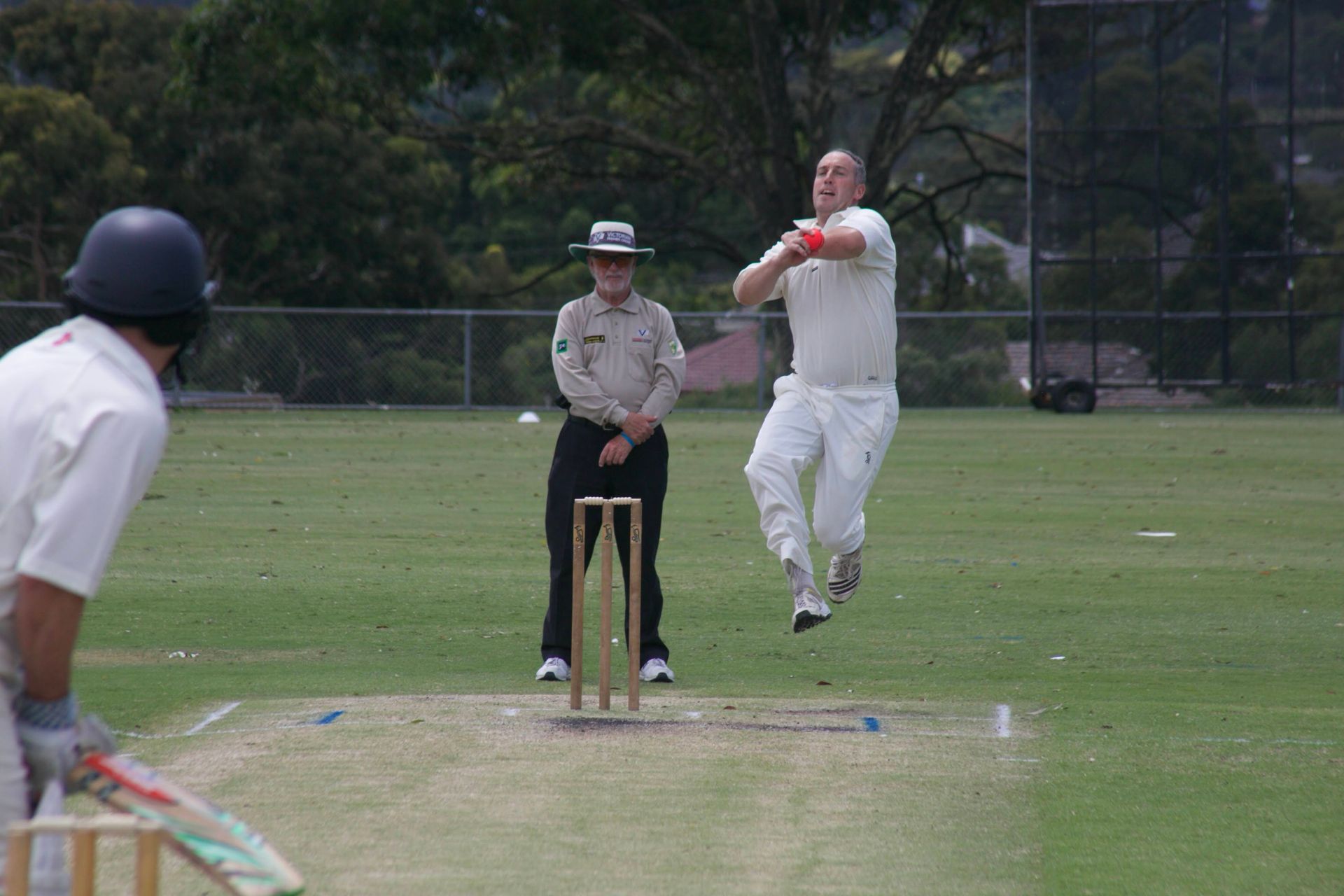 Cricket batter in white leaps to hit the ball as umpire watches behind the stumps.