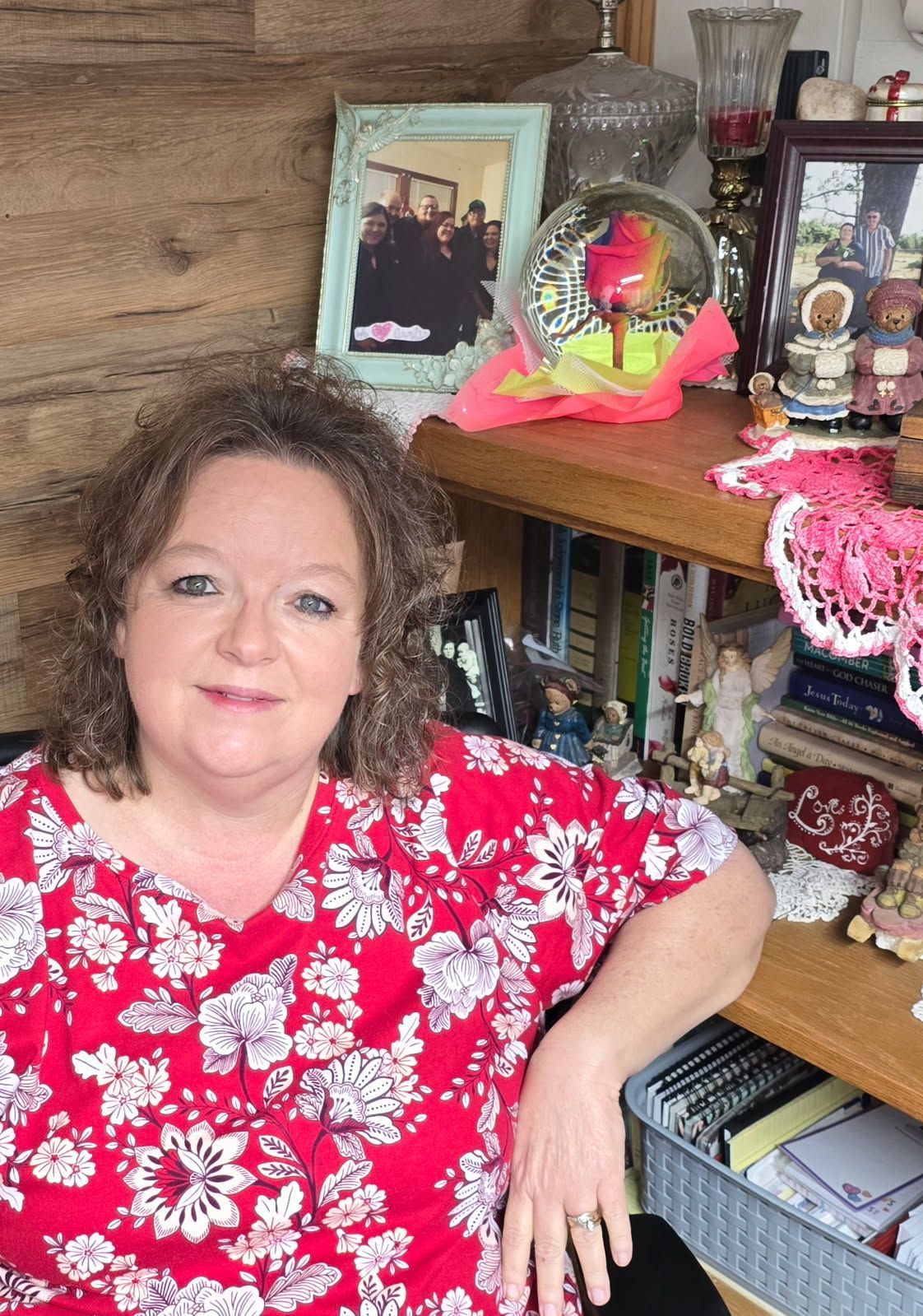 A woman in a red floral shirt is sitting in front of a shelf with pictures on it.