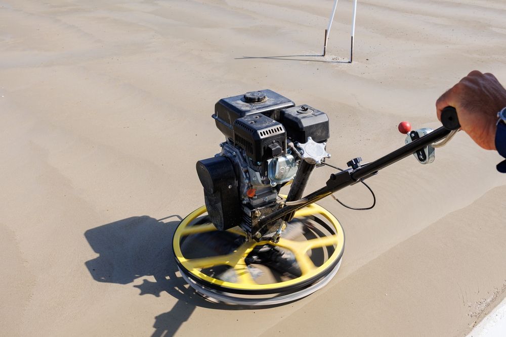 Handheld floor grinder smoothing a concrete surface, with a yellow base and operator’s hand visible.