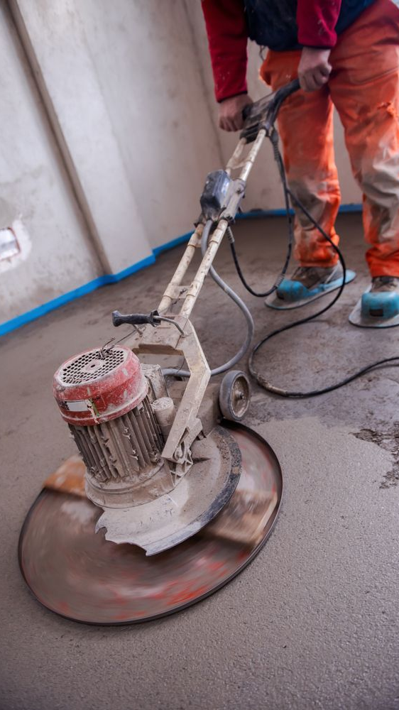Worker using a floor grinder on wet concrete indoors, with dust hose and cables visible.