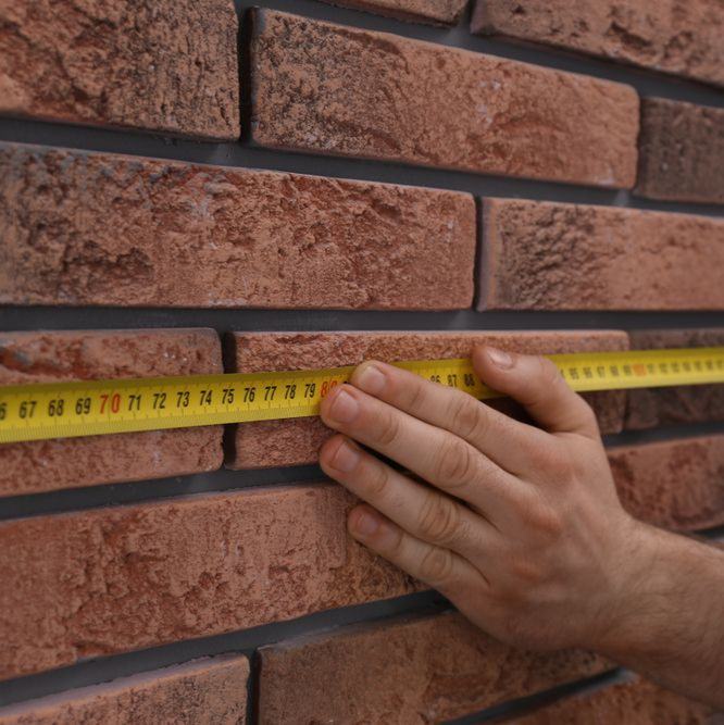 Hand measuring the width of a red brick wall with a yellow tape measure.