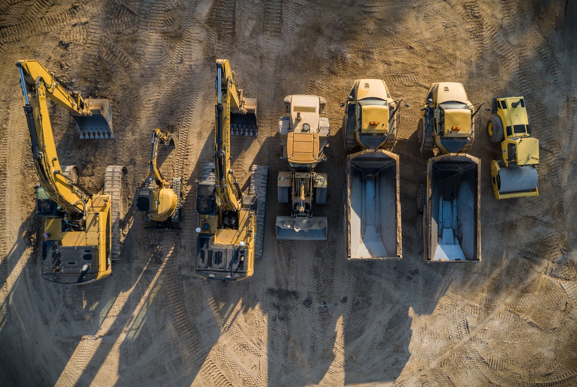 Excavators, loaders, dump trucks, and a roller lined up on a dirt construction site.