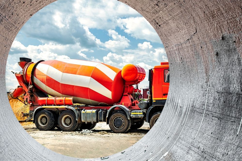 Orange concrete mixer truck viewed through a large concrete pipe at jobsite.