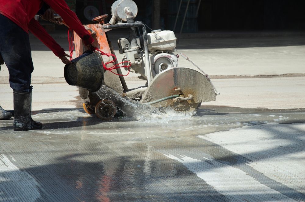 Worker using a concrete saw to cut pavement, with water spraying over the wet surface.