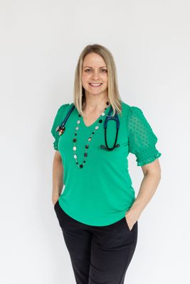Woman with stethoscope around neck, wearing green top and black pants, smiling, hands in pockets.