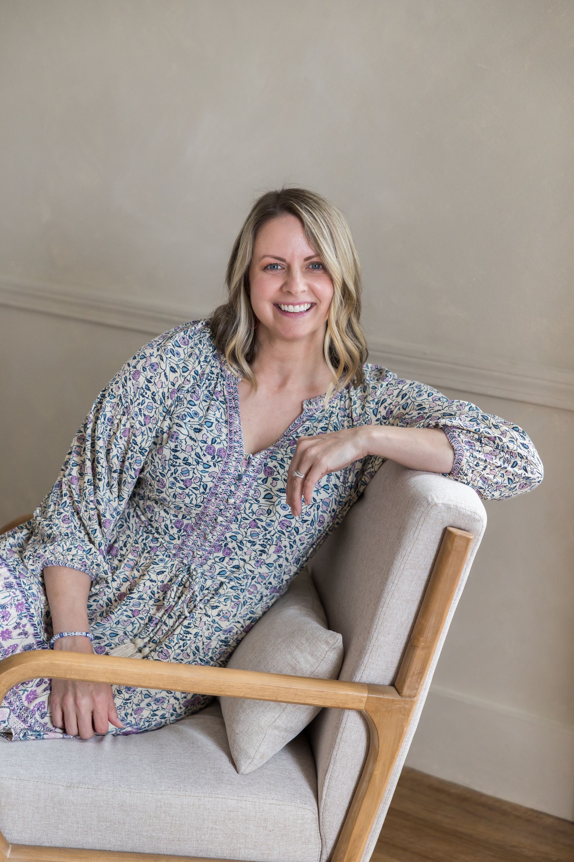 Woman in patterned dress smiles, sitting in a light-colored chair. Interior setting.