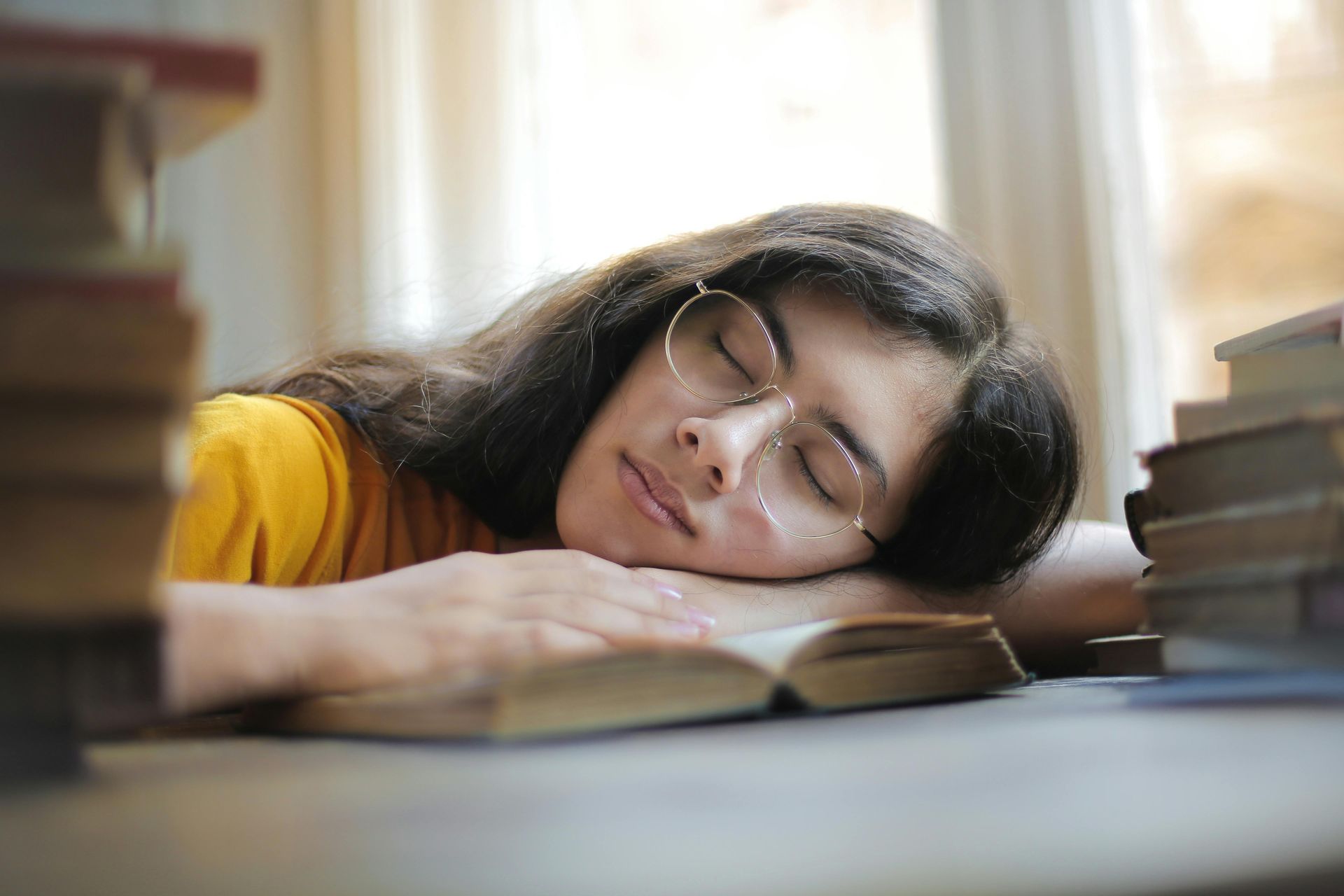 Woman with glasses asleep on an open book, resting on a table surrounded by books.