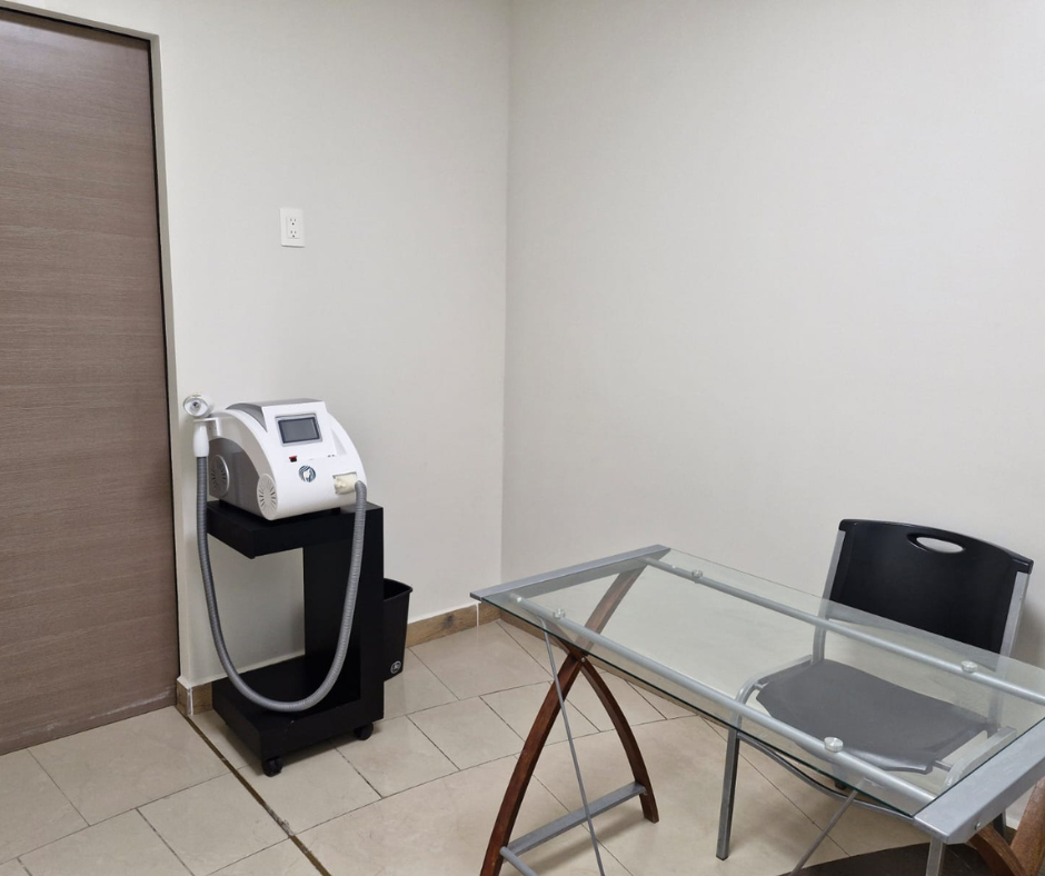 Sala médica con máquina láser, mesa de cristal y silla. Paredes beige y suelo de baldosas.