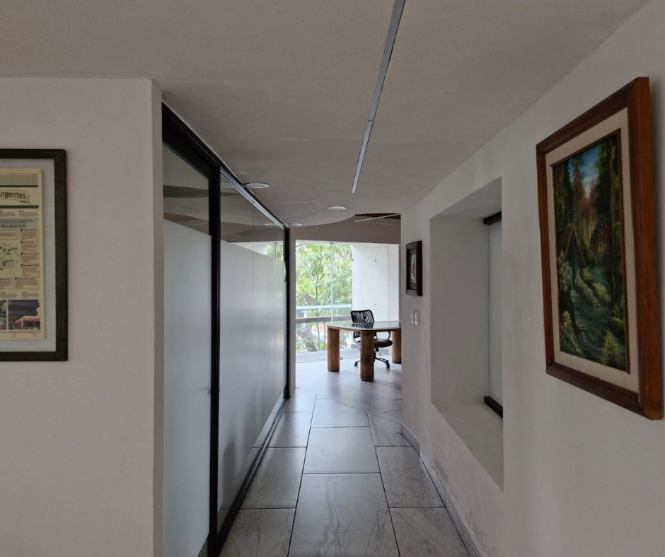 Pasillo con pared de vidrio esmerilado, suelo de baldosas grises y obras de arte. Escritorio y ventana al fondo.