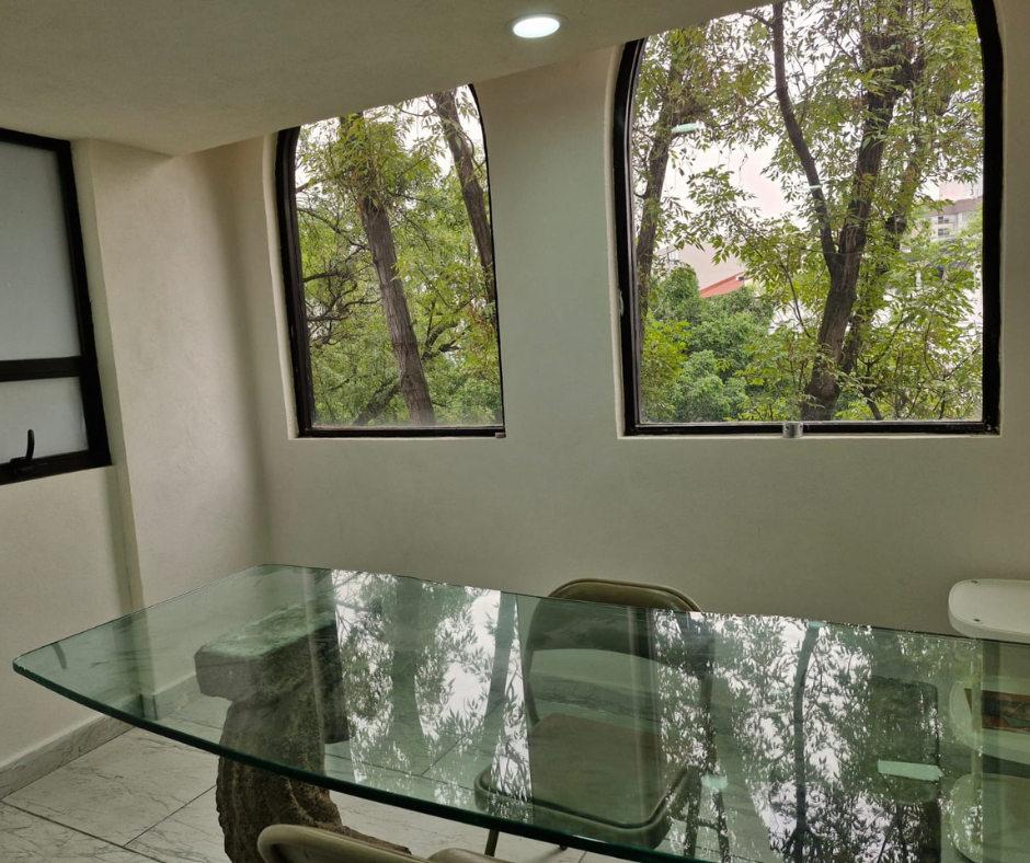 Una habitación con una mesa de cristal, dos ventanas que muestran árboles y paredes blancas.