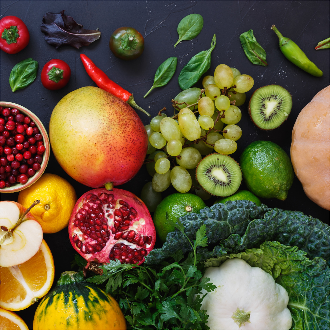 Frutas y verduras coloridas dispuestas sobre una superficie oscura, incluidos mangos, uvas, kiwis y pimientos.