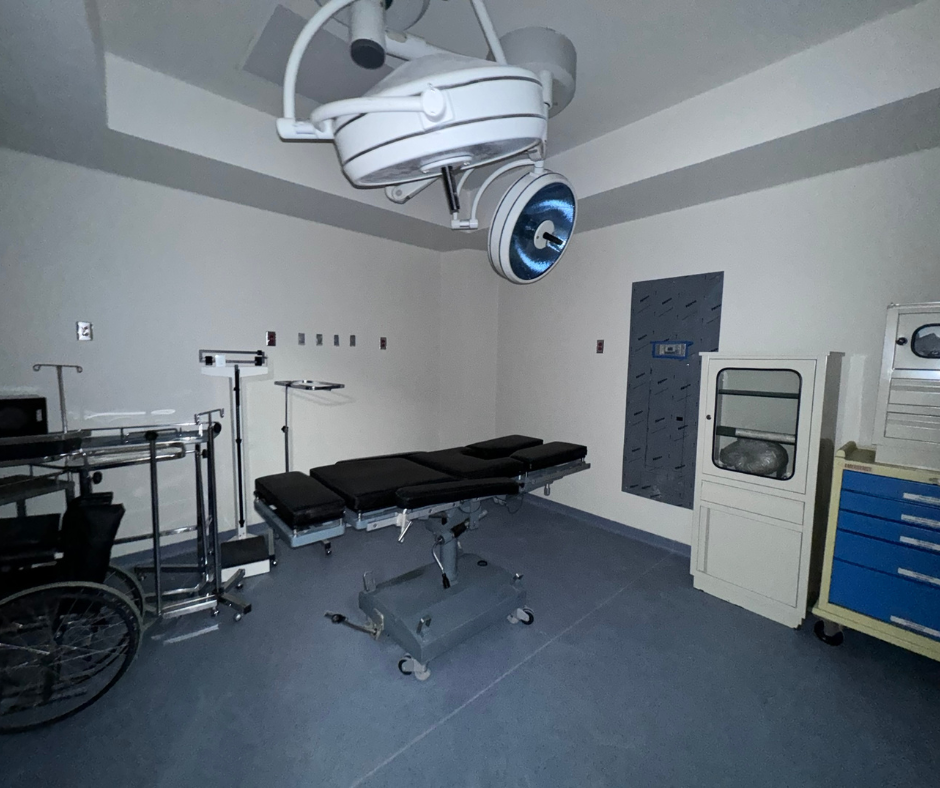 Quirófano con mesa de operaciones, lámparas de techo y equipo médico. Suelo azul, paredes blancas.