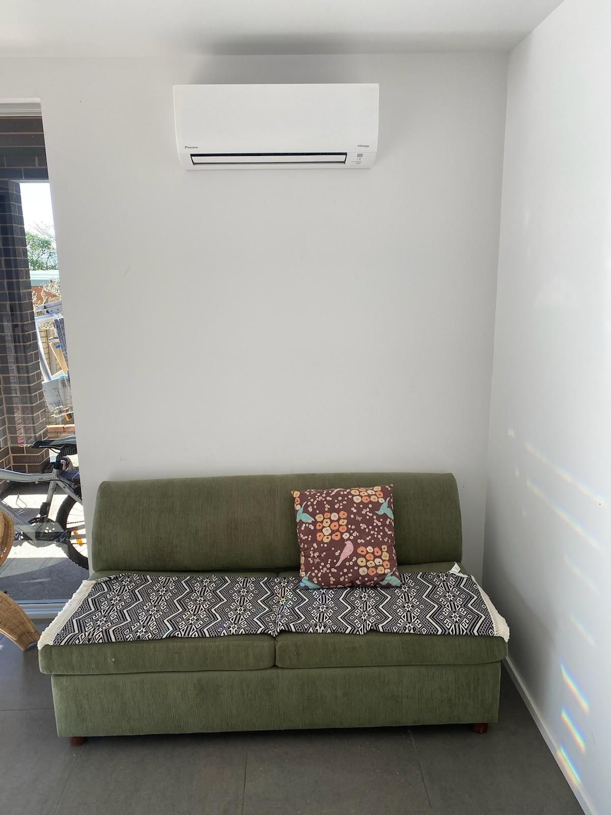 Green Sofa with Patterned Cushion, Below a White Air Conditioner  — Refrigeration & Air Conditioning in Central Coast, NSW