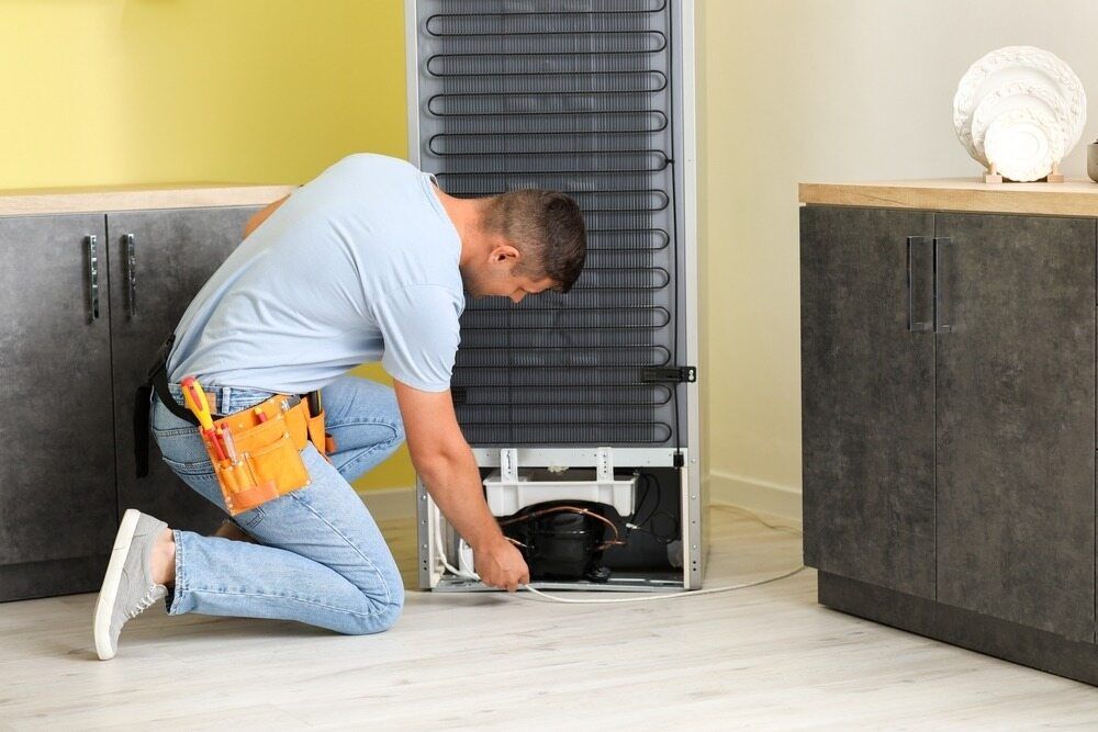 Man Kneels to Repair Refrigerator Compressor in a Kitchen — Refrigeration & Air Conditioning in Warners Bay, NSW