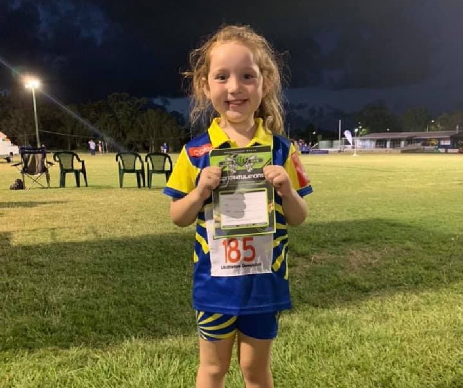 Girl in blue and yellow sports uniform holding a Laserzone Sunshine Coast achievement award on a grass field at night.