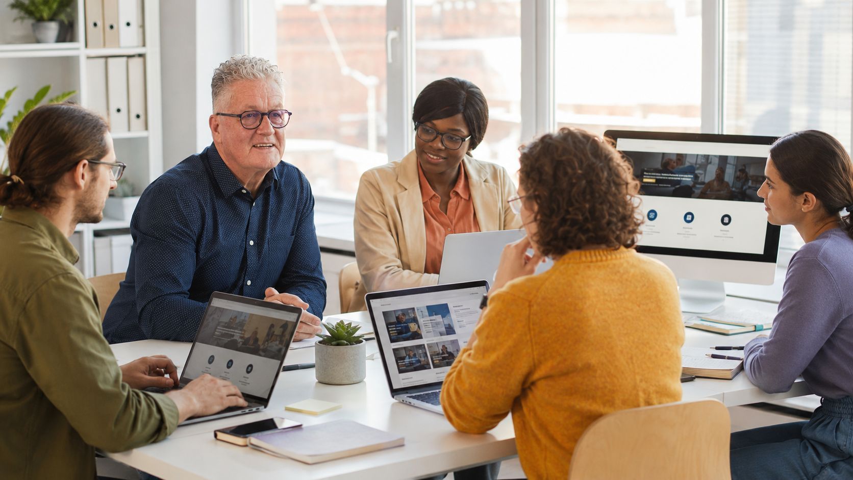 People collaborating in a bright office; some working on laptops and others discussing documents near a window.