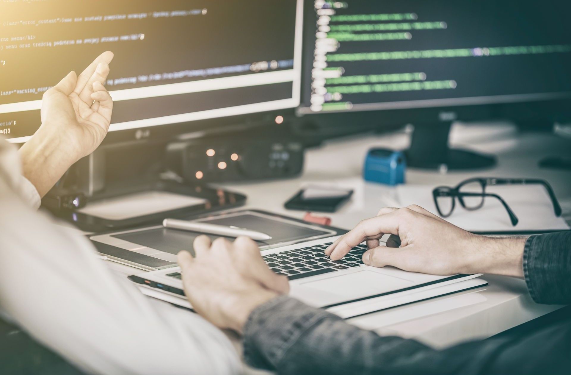 Two people working on computers with code, one pointing and typing, indoors.