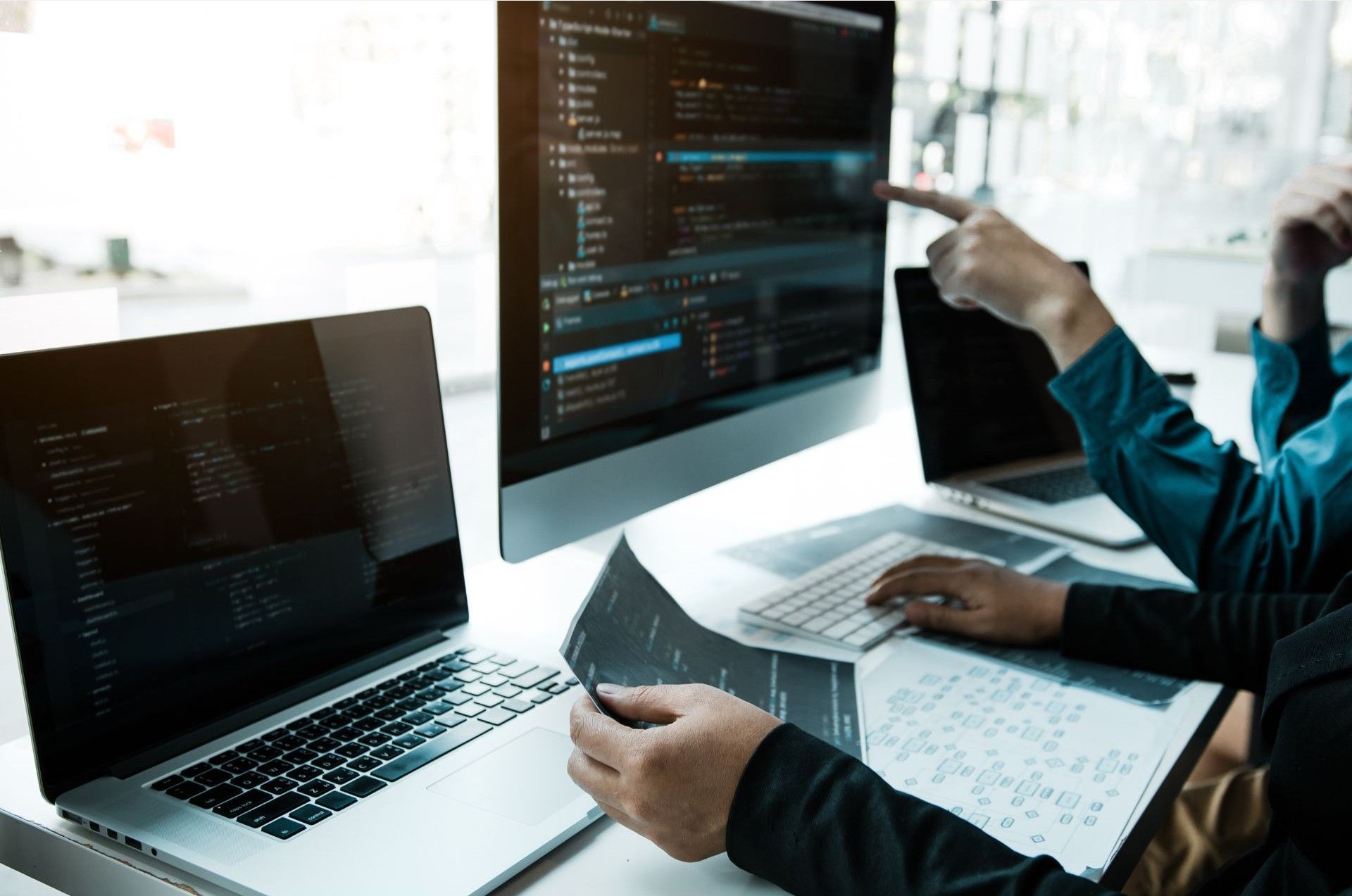 Two people reviewing code on computer monitors and printouts, lit by natural light.