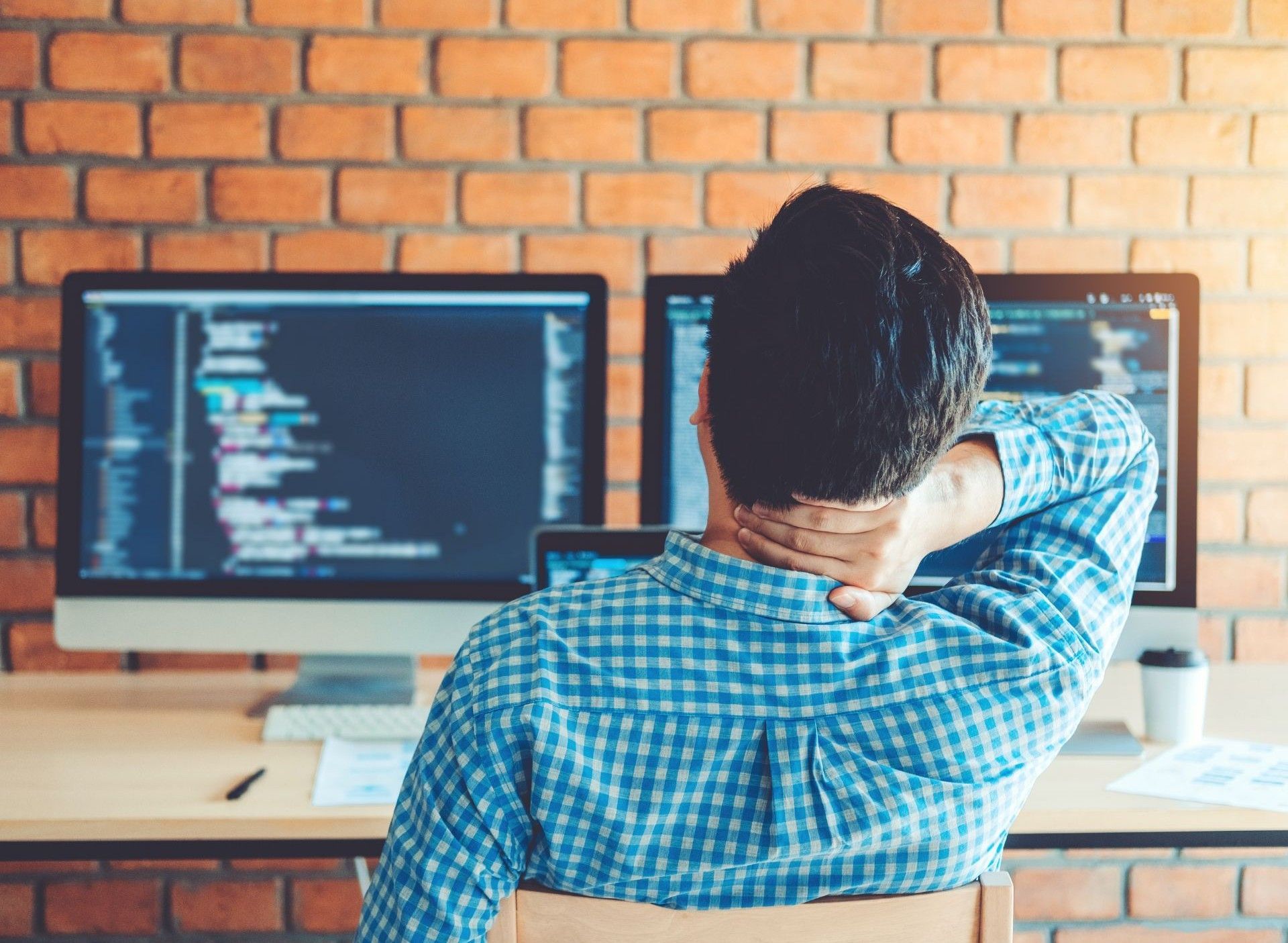 Man at desk with neck pain, facing two computer monitors displaying code, brick wall backdrop.