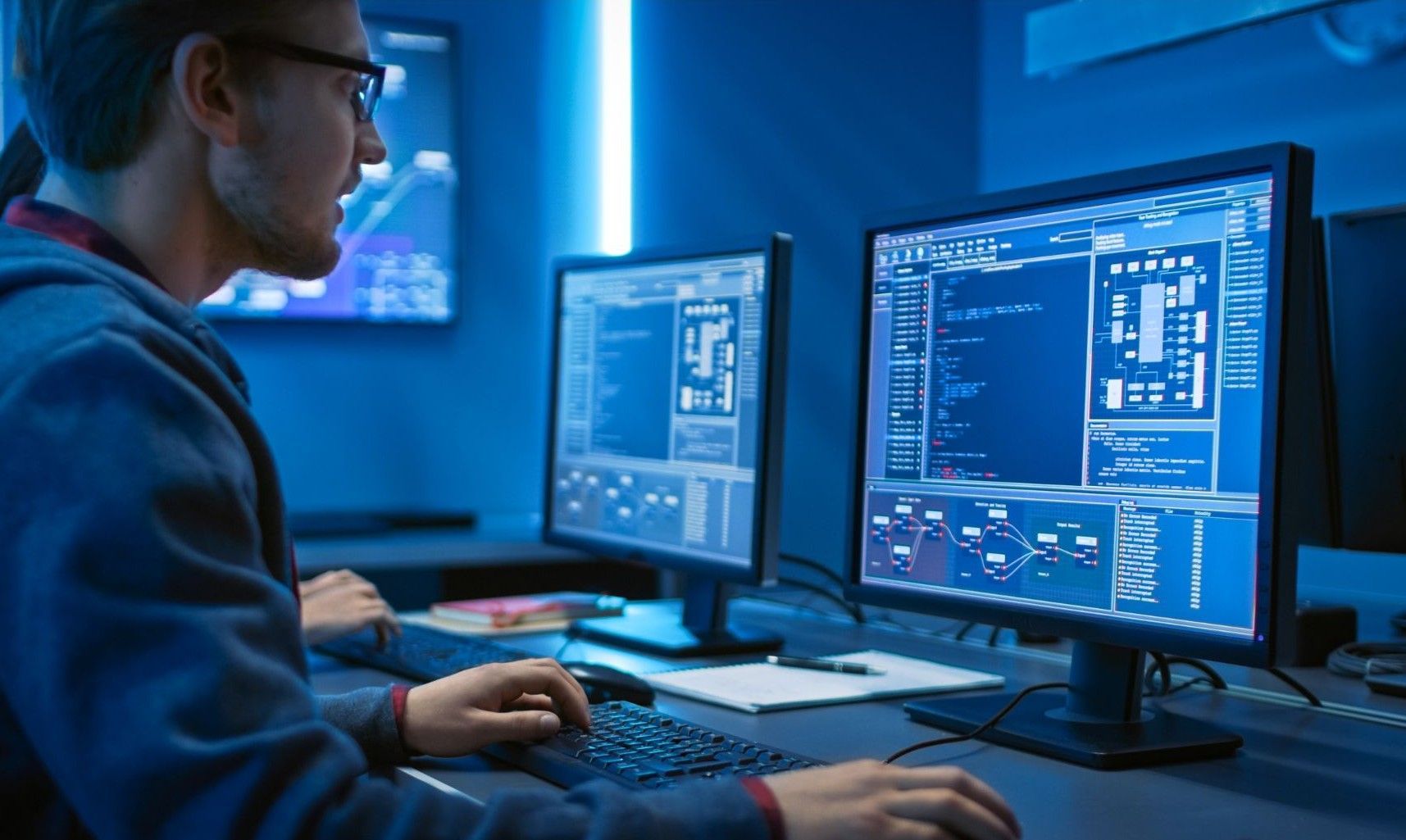 Man seated at computer, coding; blue-lit room, two monitors displaying code.