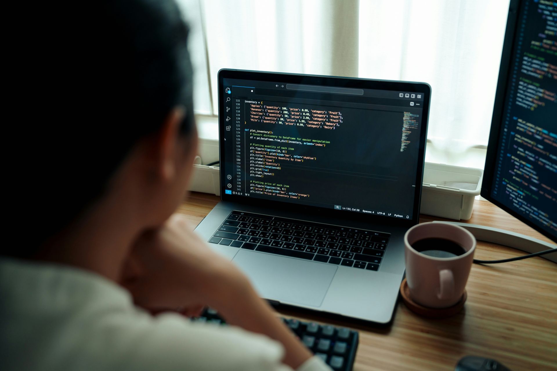 Person looking at laptop with code on the screen and a second monitor. Coffee mug on desk.
