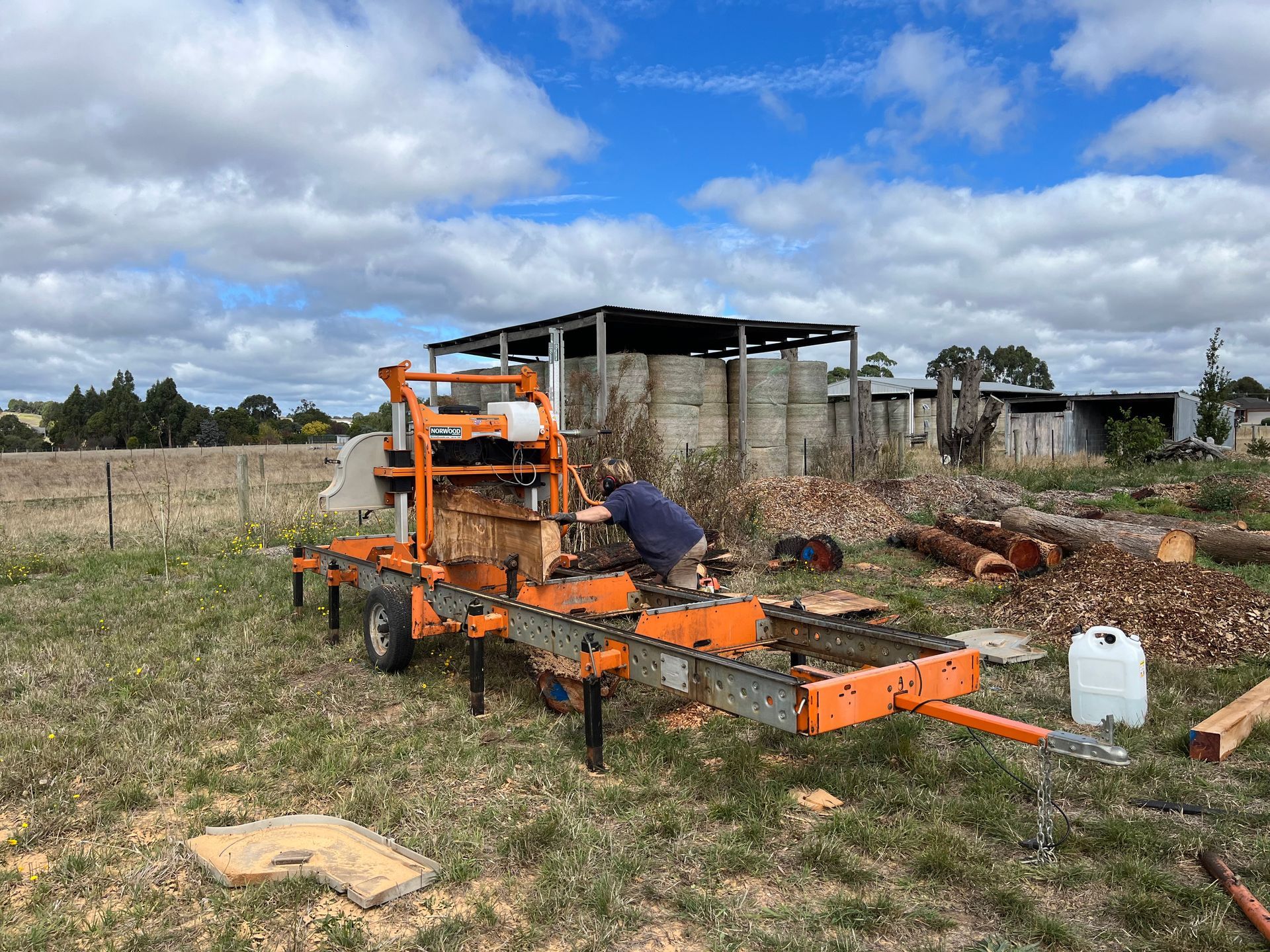 Person sawing a log on an orange portable sawmill in a field with wooden structures in the background.