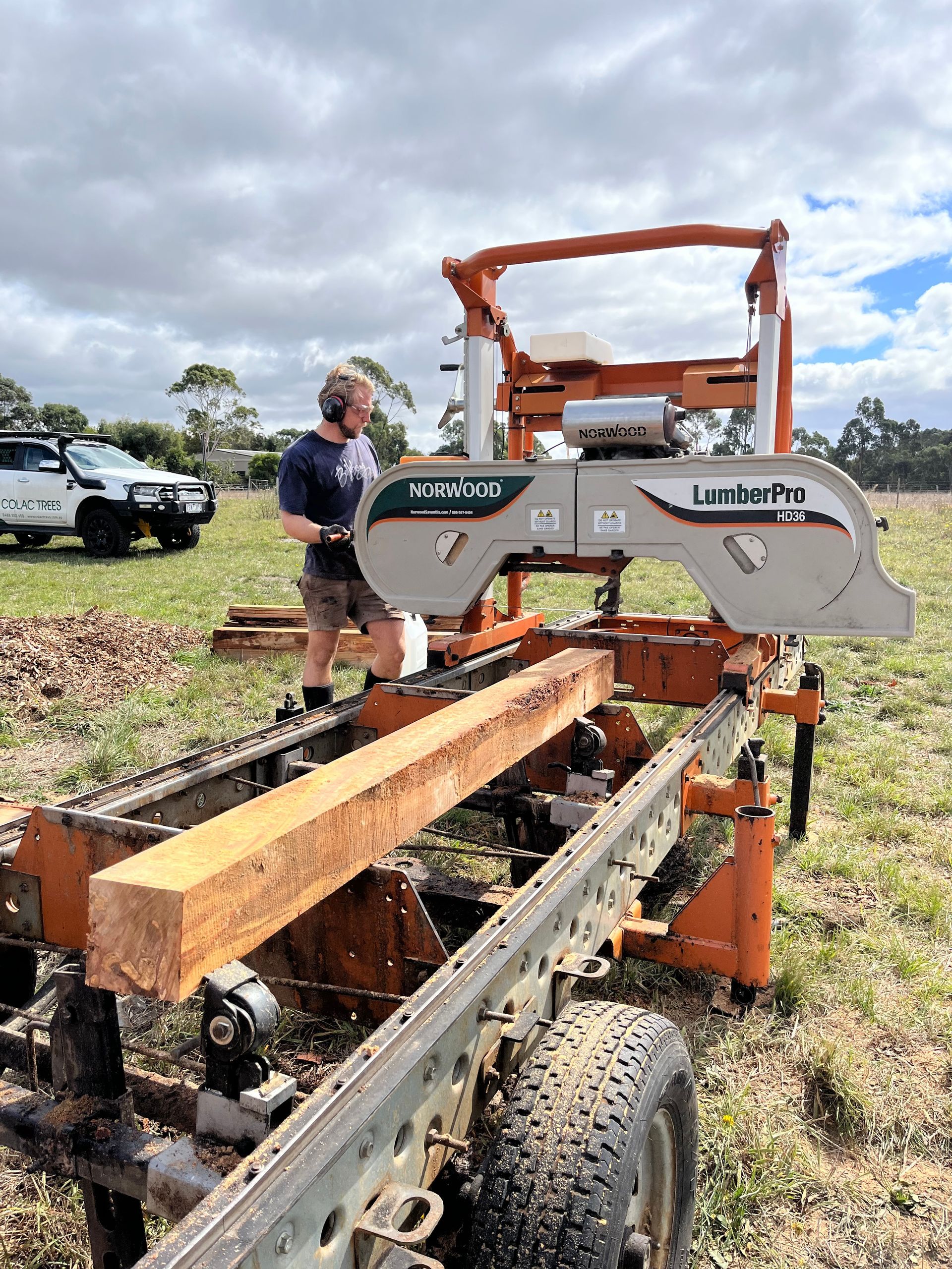 Man operating a portable sawmill outdoors, cutting a large wood beam on a trailer. Cloudy sky.