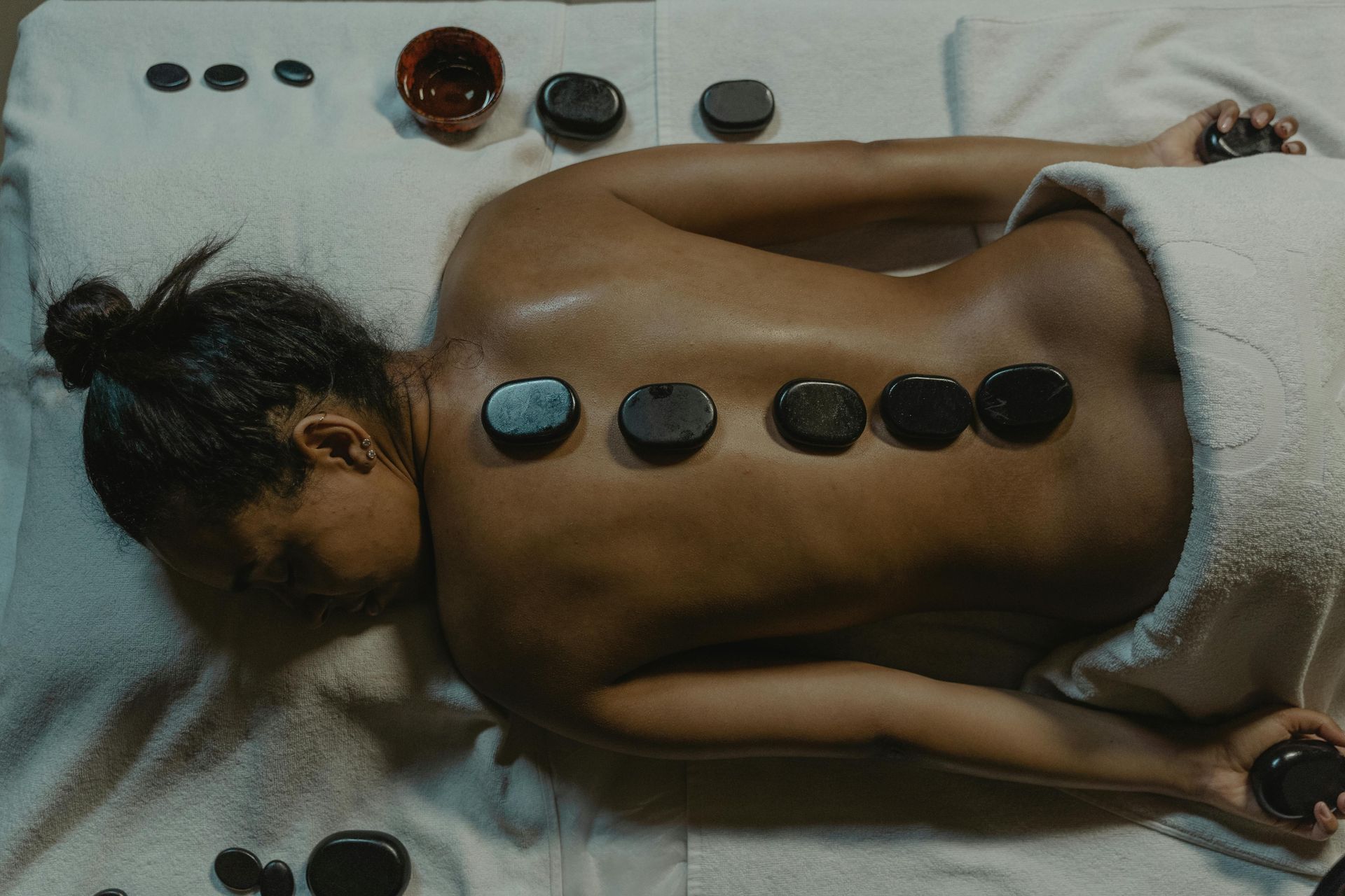 Person receiving a hot stone massage, therapist placing stones on back. Candles in background.