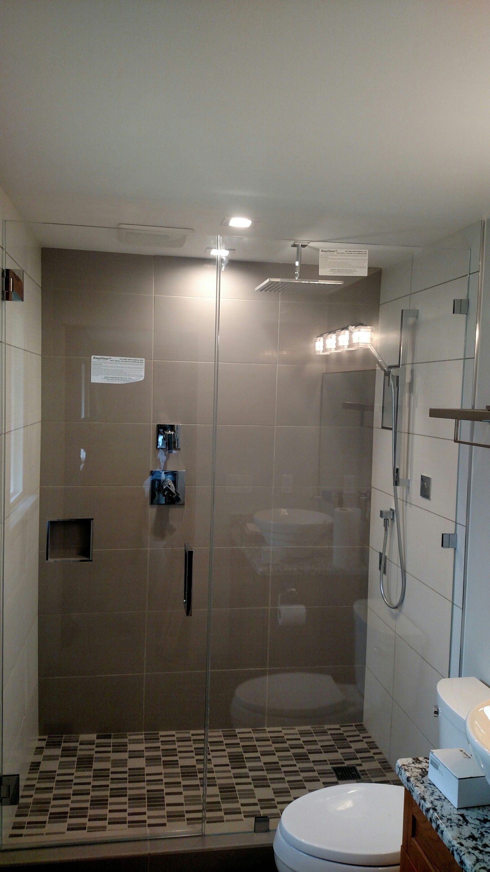 Bathroom with a walk-in shower. Gray tiled walls, glass doors, and a pebble-stone floor. White toilet in front.