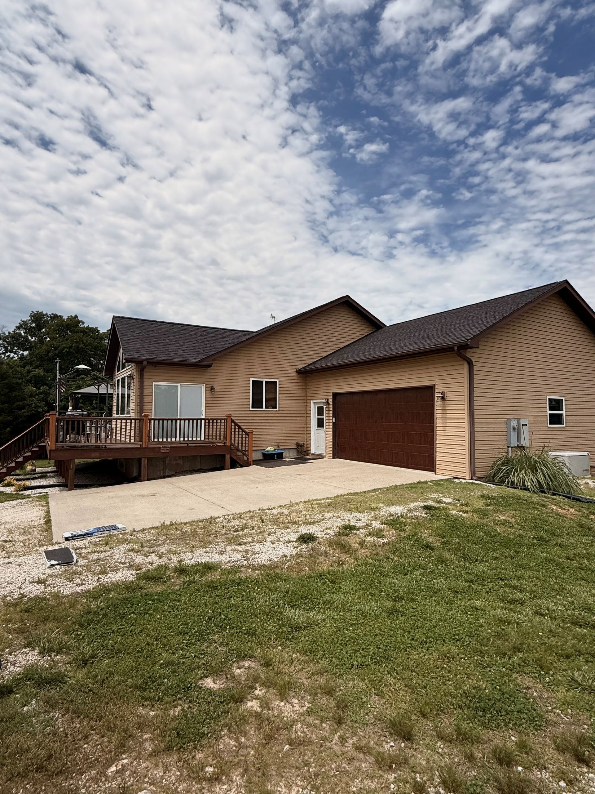 House with tan siding, brown roof, attached garage, and deck under a cloudy sky.