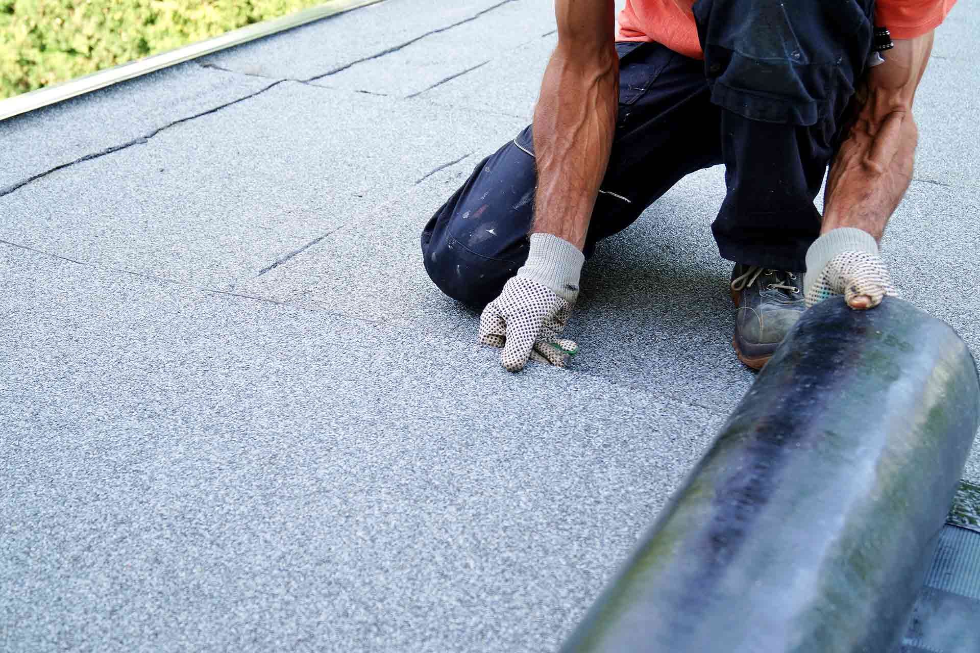 Man installing a rolled roofing material on a flat roof, wearing gloves and dark pants.
