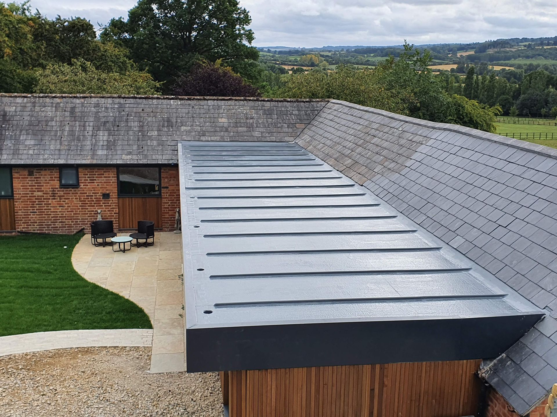 A modern flat metal roof extension attached to a traditional brick and slate building overlooking a rural landscape.