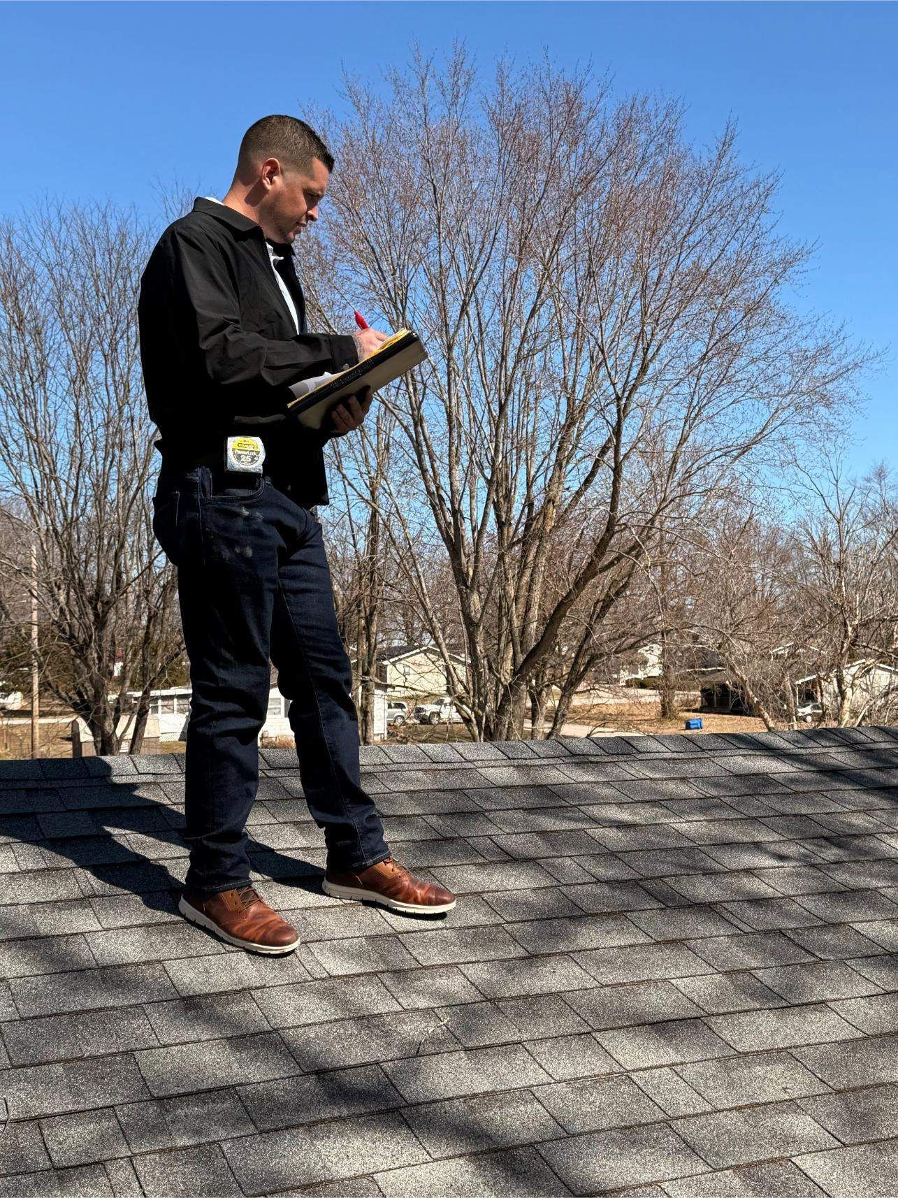 A person in dark work clothes standing on a shingled roof, writing on a clipboard under a clear blue sky.