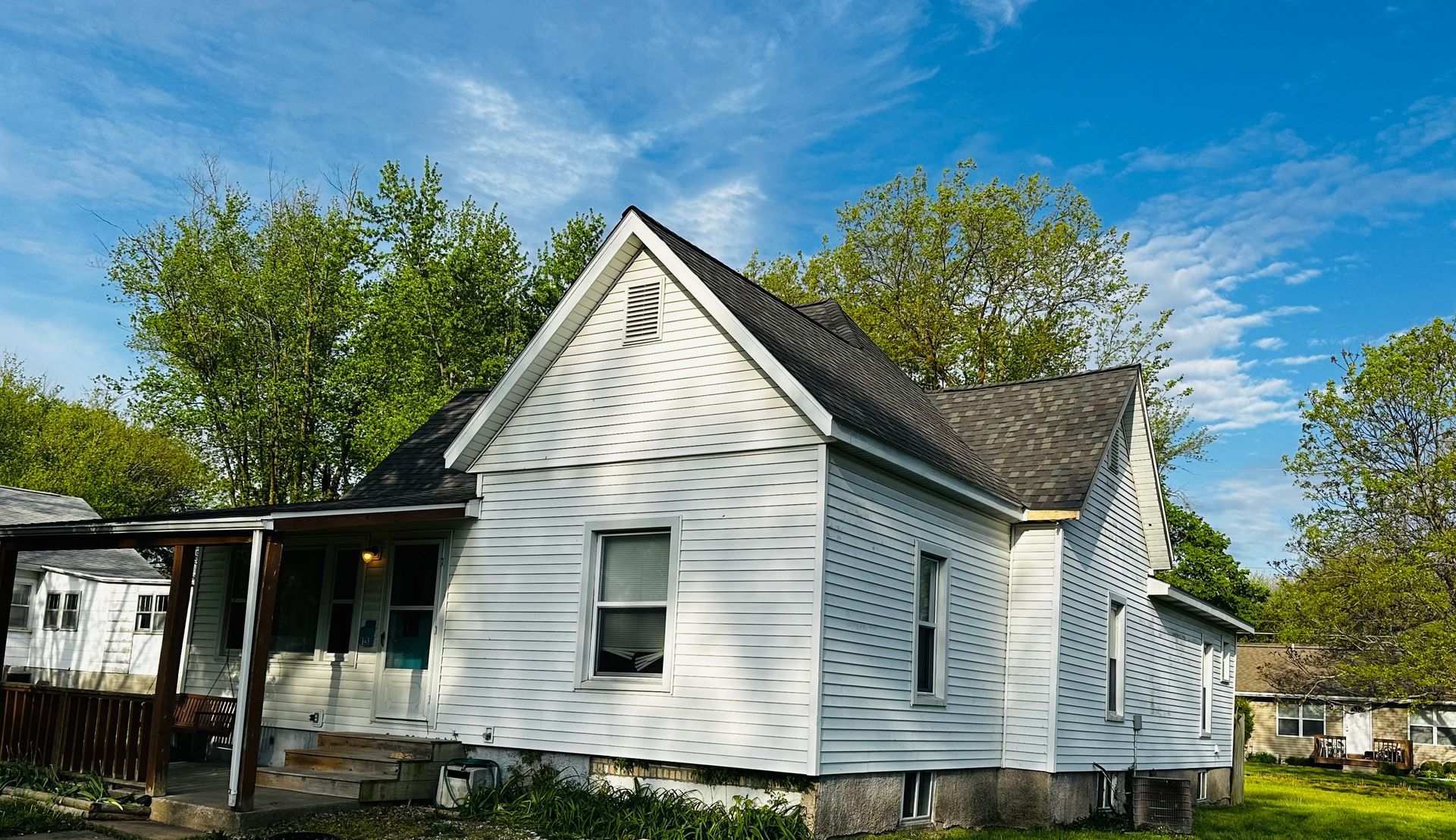 White, one-story house with a porch and dark roof against a blue sky with trees.
