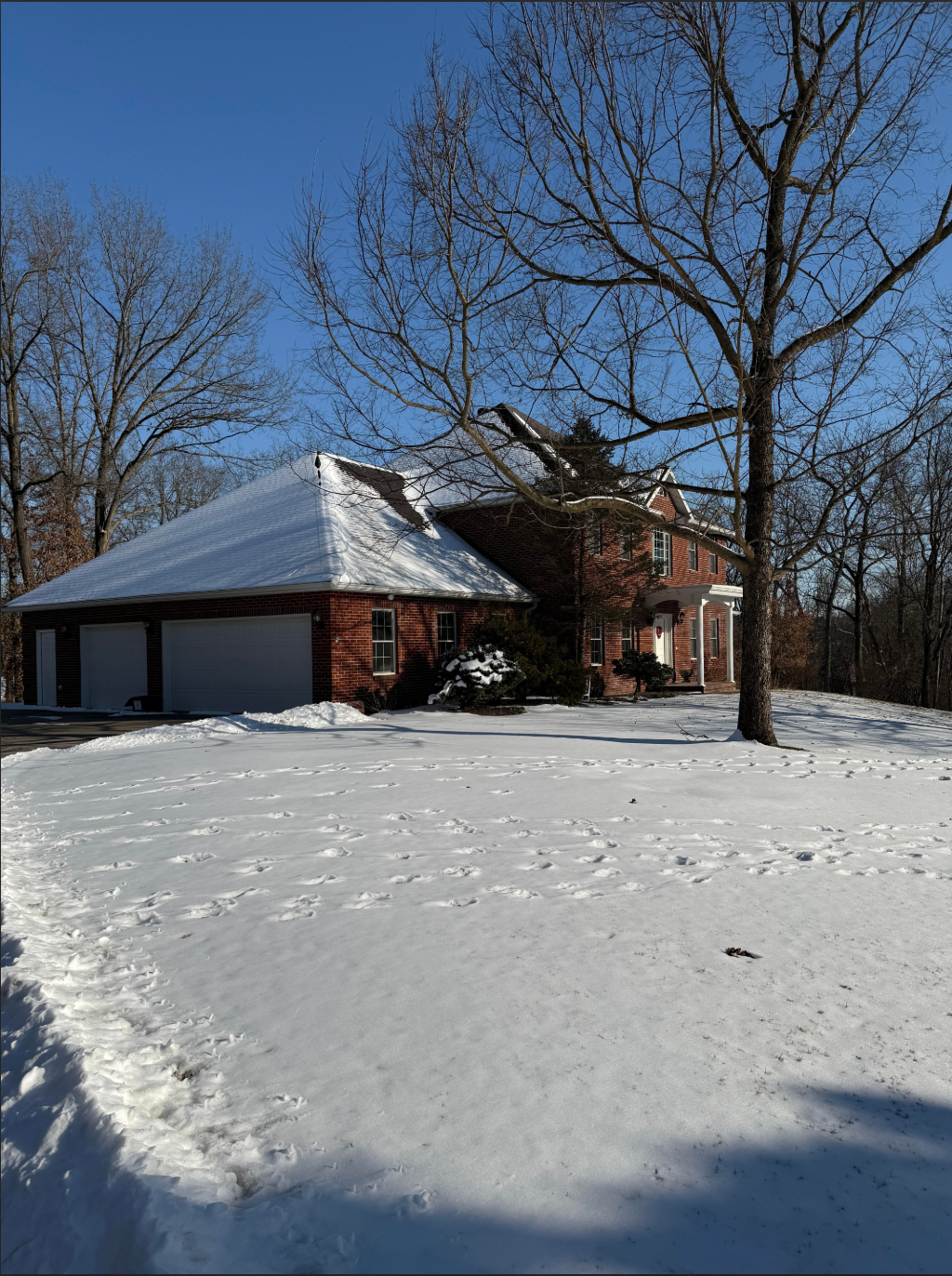 Snow-covered house with attached garage, red brick, and bare trees under a blue sky.