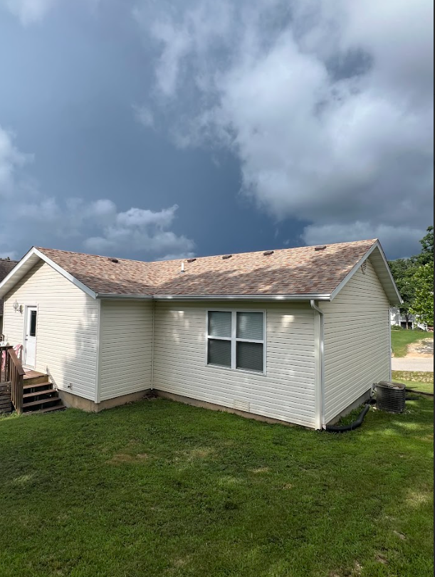 A beige house with a brown roof under a stormy sky. Green grass in the yard.