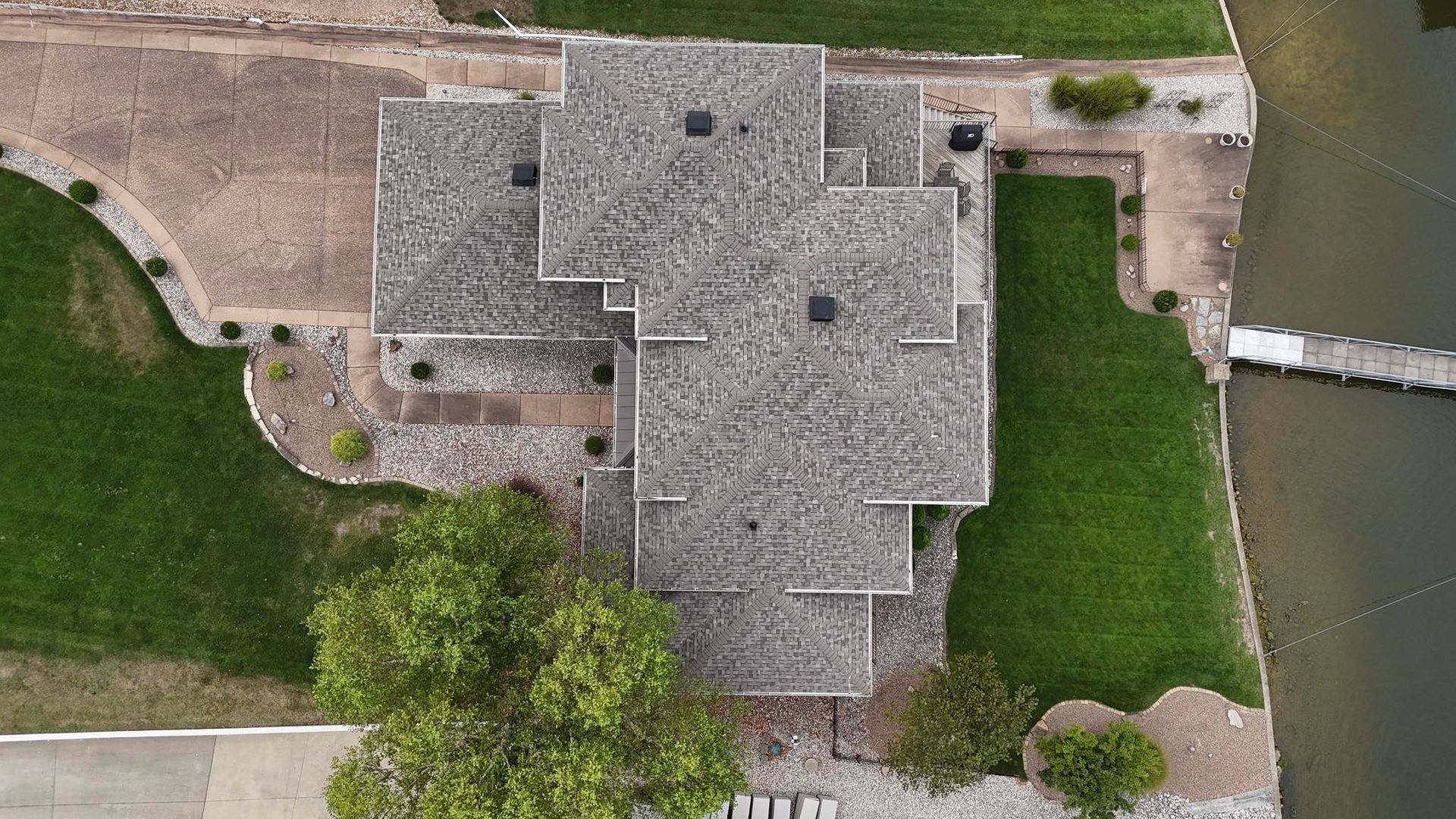 Aerial view of a gray-roofed house with green lawn, next to a body of water and a dock.