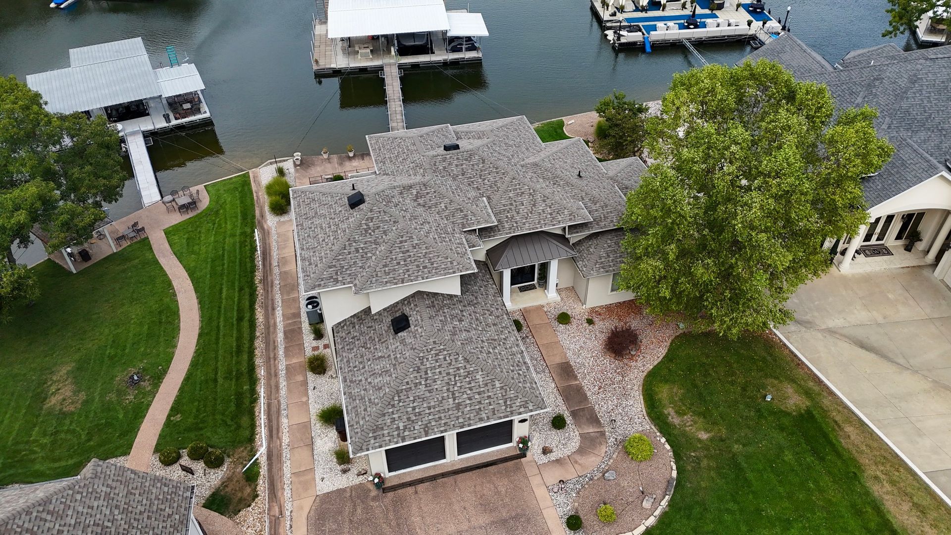 Aerial view of a lakeside home with a gray roof and attached garage, surrounded by green grass and docks.