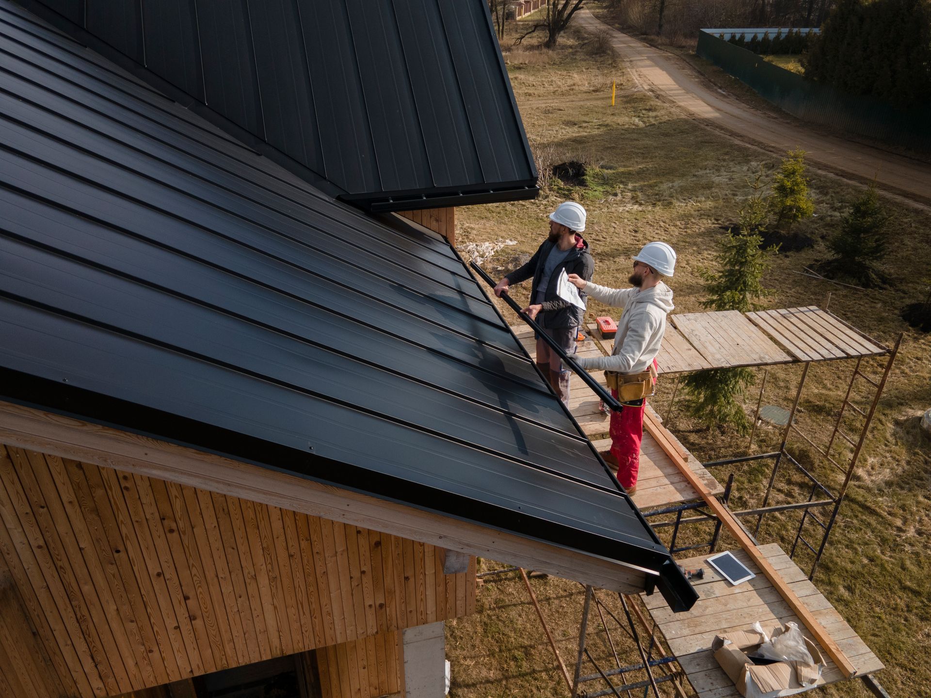 Two roofers on a dark metal roof, using tools and standing on scaffolding.