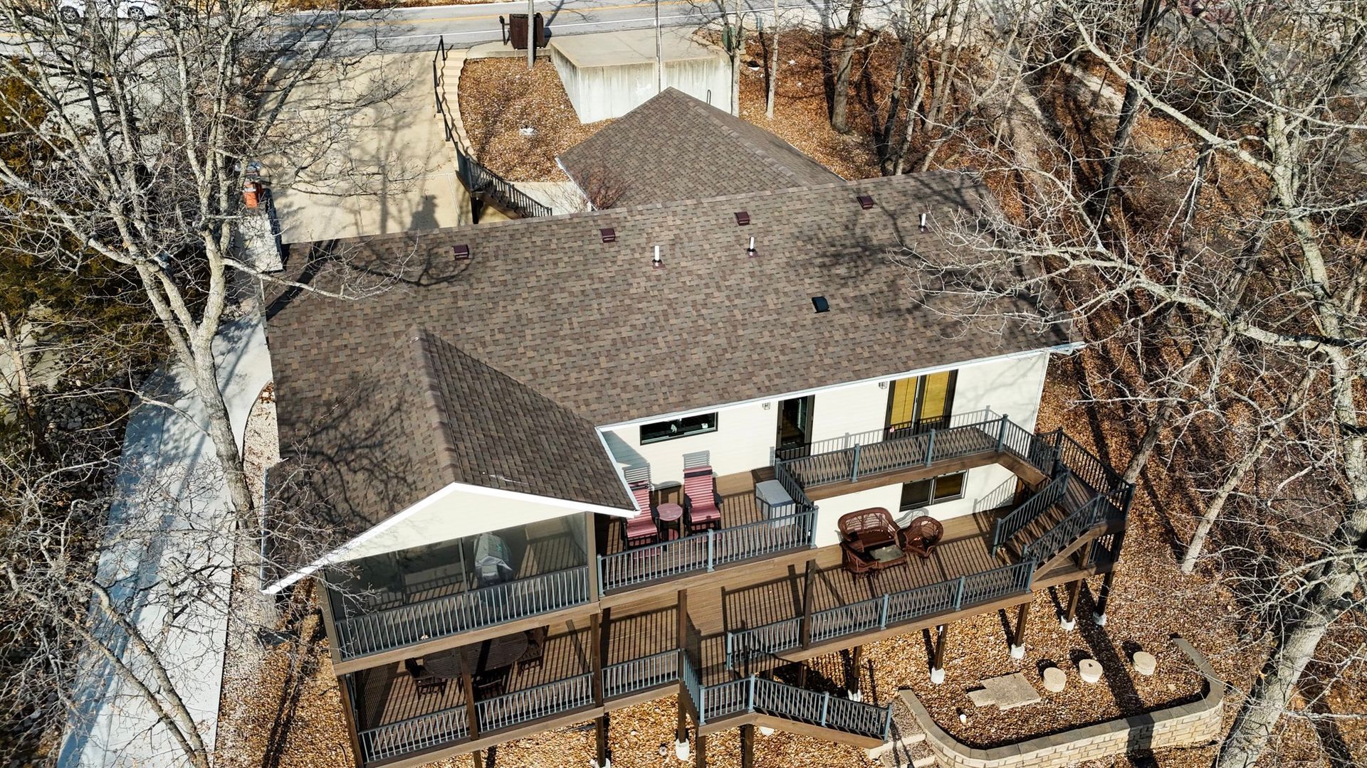 Aerial view of a white single-story house with a large wooden deck, nestled among bare trees in a wooded setting.