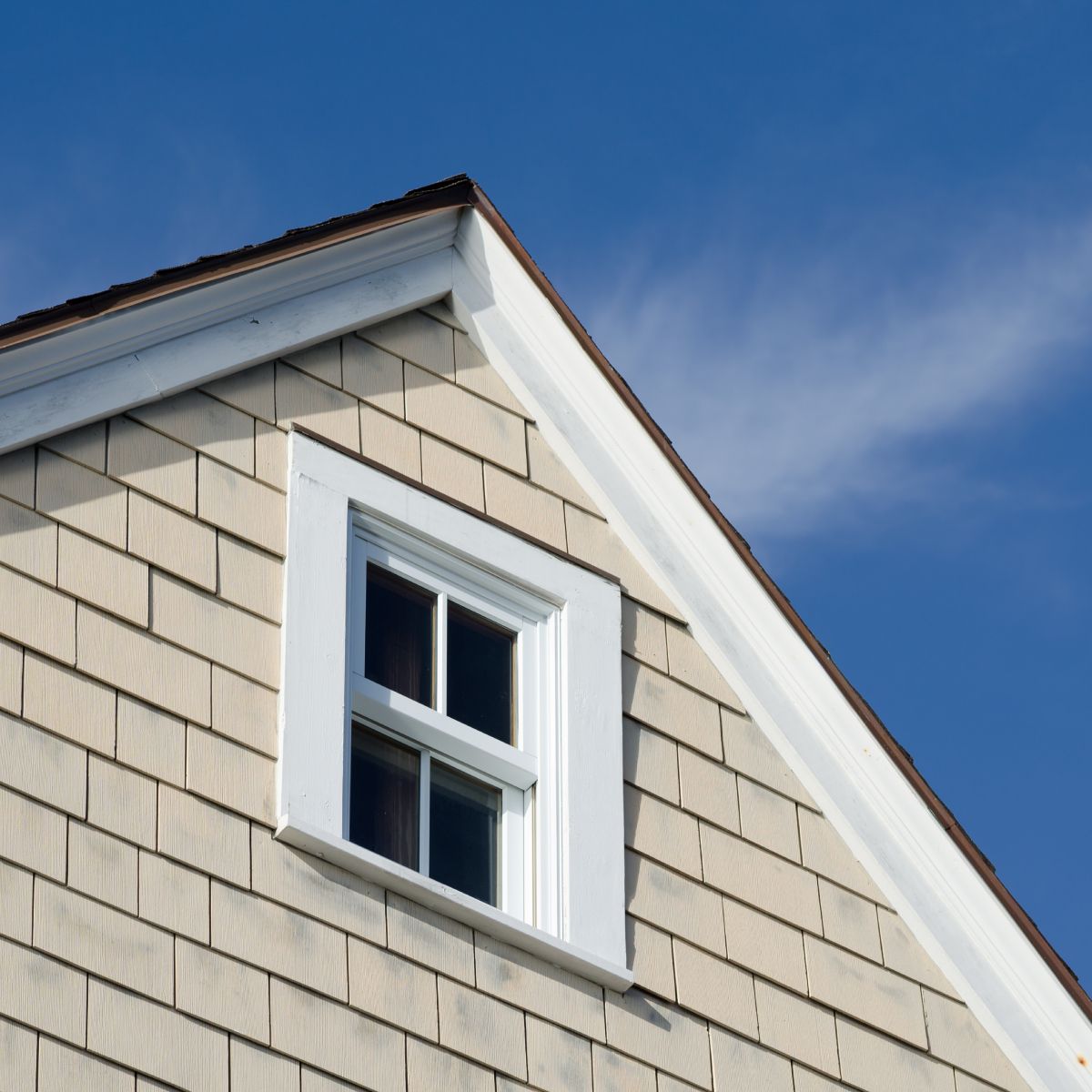 Gabled house exterior with shingle siding, a white window frame, and blue sky.