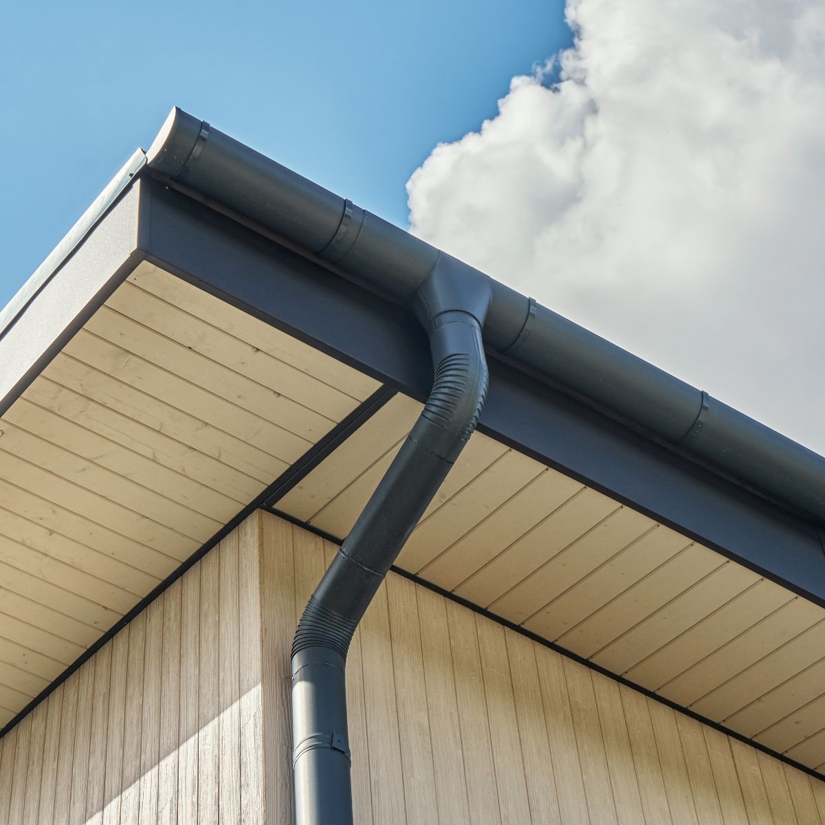 Black gutter system on a modern building's corner with wood-look siding. Blue sky and clouds above.