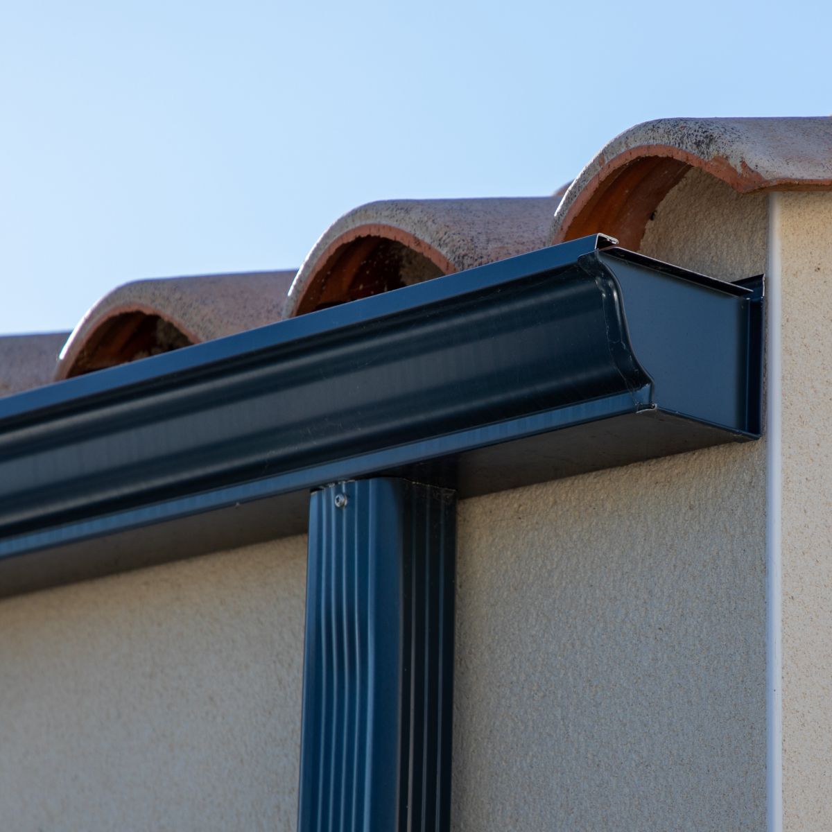 Black rain gutter along a tan stucco wall, under terracotta roof tiles.
