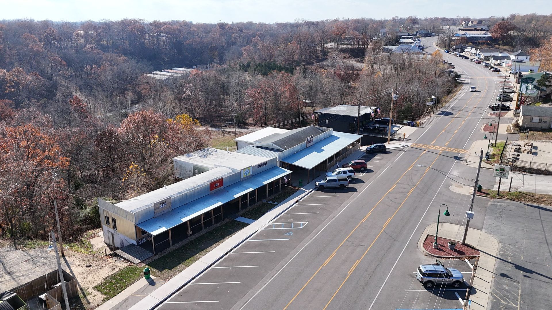 Aerial view of a long, single-story building with a covered porch and parking along a street lined with trees and other buildings.
