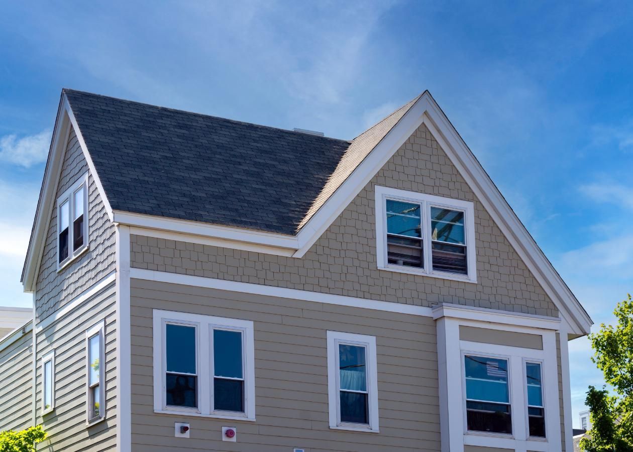 Tan and gray two-story house with gabled roof, blue windows, and a blue sky background.