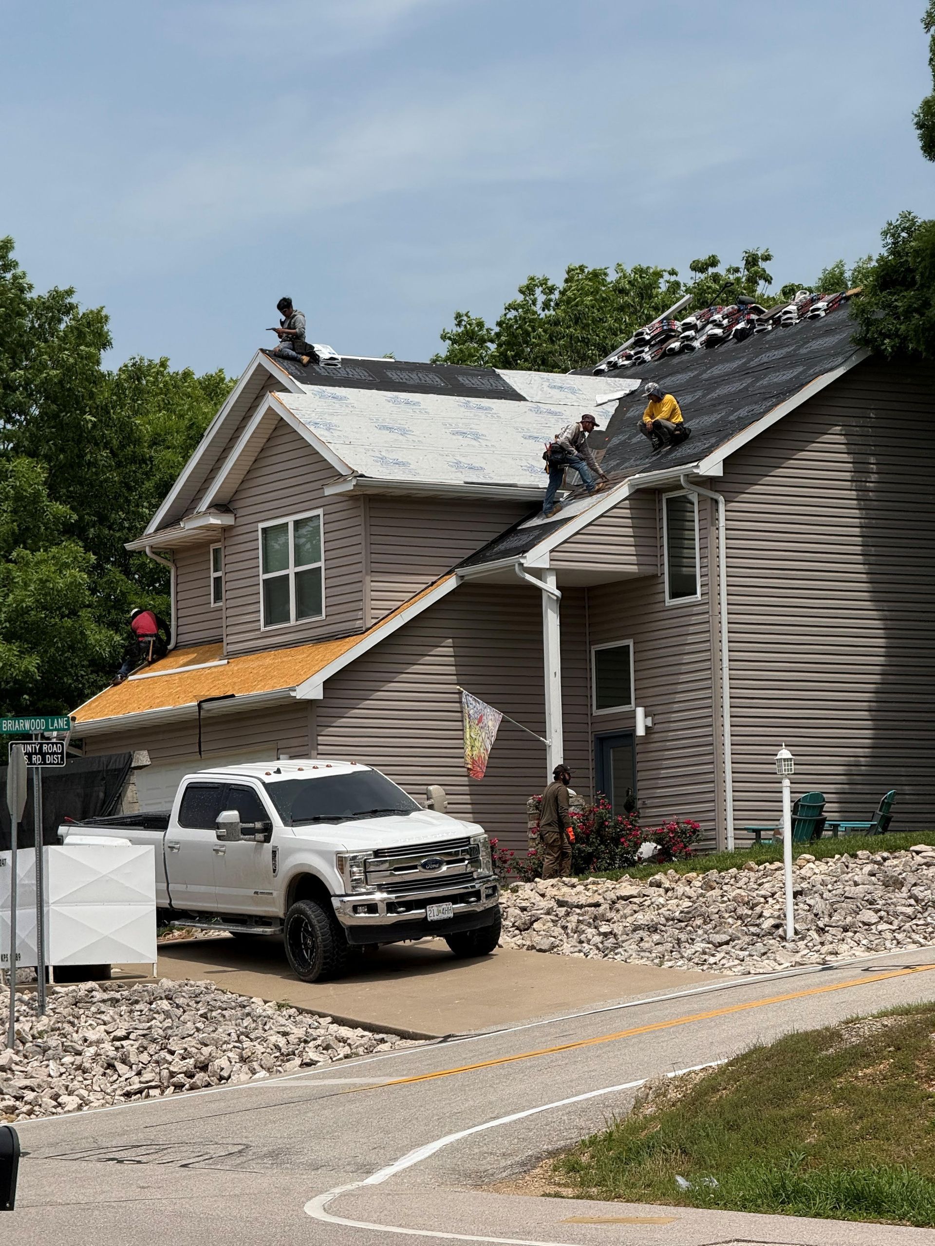 Workers on roof replacing shingles; white truck parked in front of two-story house, sunny day.